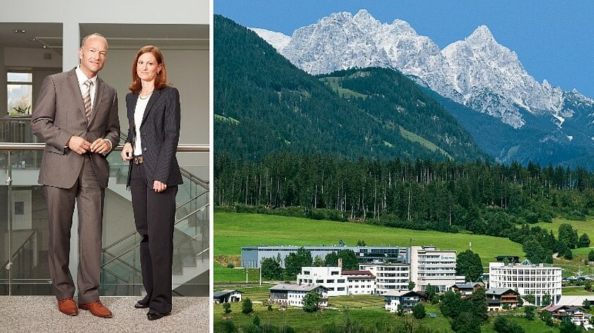 Ein Mann und eine Frau stehen neben einer Panoramaaufnahme eines Firmengeländes, im Hintergrund grüne Wiesen, Wald und hohe Berge