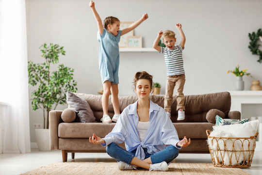 Zwei aufgedrehte Kinder hüpfen auf einer Couch, Mutter sitzt am Boden und meditiert